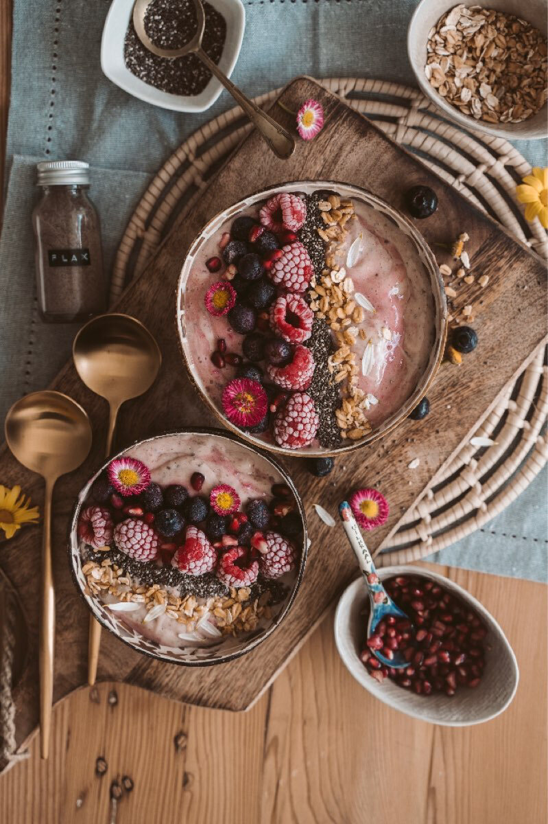 Healthy smoothie bowls sit atop wooden table with fruit, spices and golden silverware surrounding them.