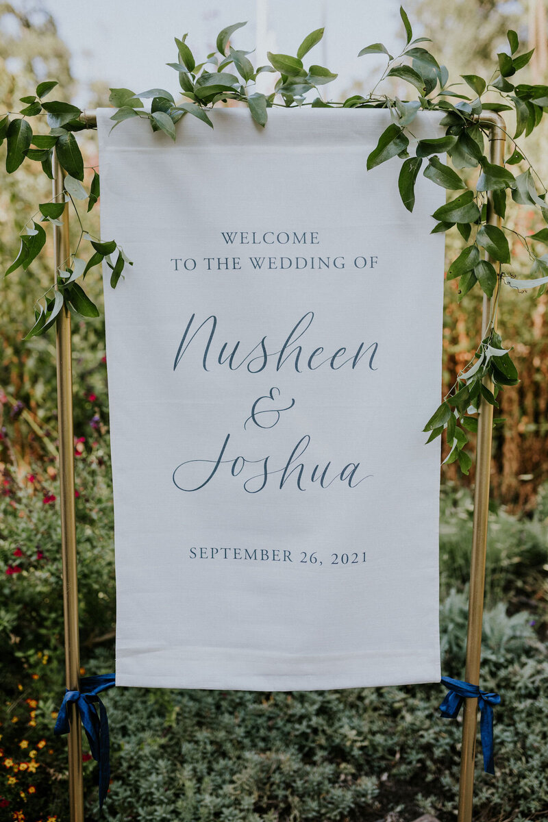 Fabric welcome sign with navy lettering and ribbon, hanging on a gold stand
