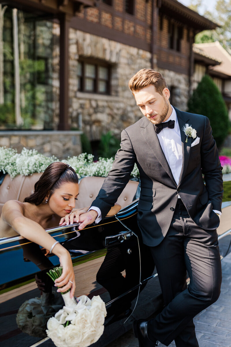 Groom in a gray tuxedo jacket leaning on a vintage car beside his bride and her white bouquet