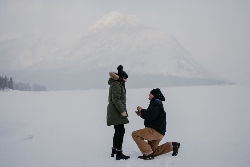 A young couple sit on driftwood on the shore of a mountain lake during their photography session. She has her hands clasped on one raised leg and is looking down and he's sitting beside her, kissing her on the side of her head.
