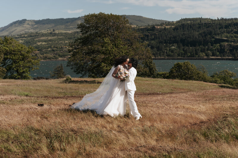 BIPOC lesbian couple in wedding attire embracing on a summer hillside overlooking the Columbia River Gorge with Mount Hood in the background, captured by an LGBTQ+ inclusive wedding photographer specializing in adventurous Pacific Northwest elopements and weddings.