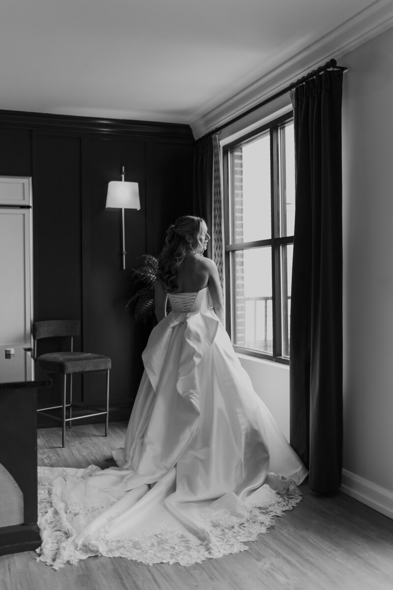 A BLACK AND WHITE  PHOTO OF A BRIDE LOOKING OUT A WINDOW 