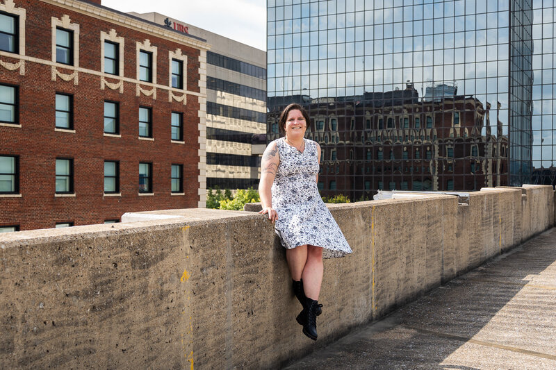 A woman sits confidently on the edge of a concrete ledge, surrounded by tall buildings and reflecting windows. She wears a patterned dress and combat boots, smiling with relaxed ease. Her tattoos peek out as she leans slightly forward, radiating authenticity and strength. It’s a moment of individuality and quiet boldness, full of presence and personal style.