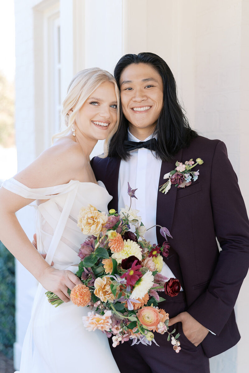A joyful newlywed couple stands smiling at Donovan Manor, the bride in an off-the-shoulder gown holding a lush, fall-toned bouquet and the groom in a plum suit with a coordinating boutonniere; this romantic wedding moment was published and featured in Magnolia Rouge and designed by Grey Cove Designs.