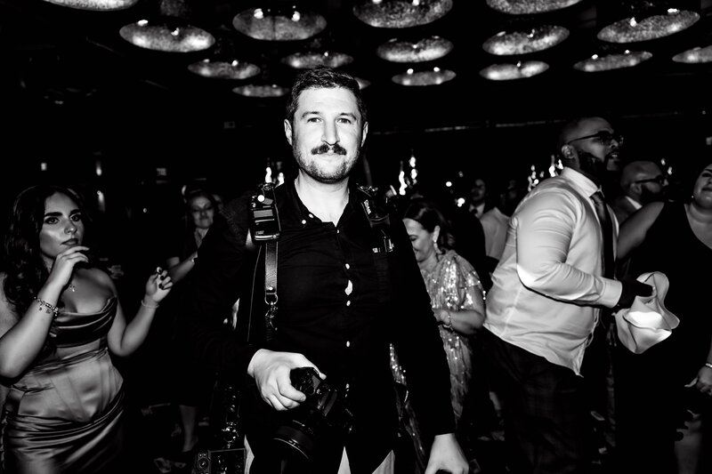 A man with a mustache, wearing a black shirt and camera strap, stands holding a camera in a crowded, dimly lit room where people are dancing and celebrating, perfectly capturing the moment as an experienced NJ wedding photographer.