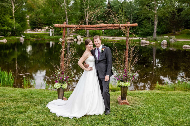 A cute couple poses for a formal wedding picture in front of their wood arch in front of the pond on the lawn area at Yampa River Botanic Park