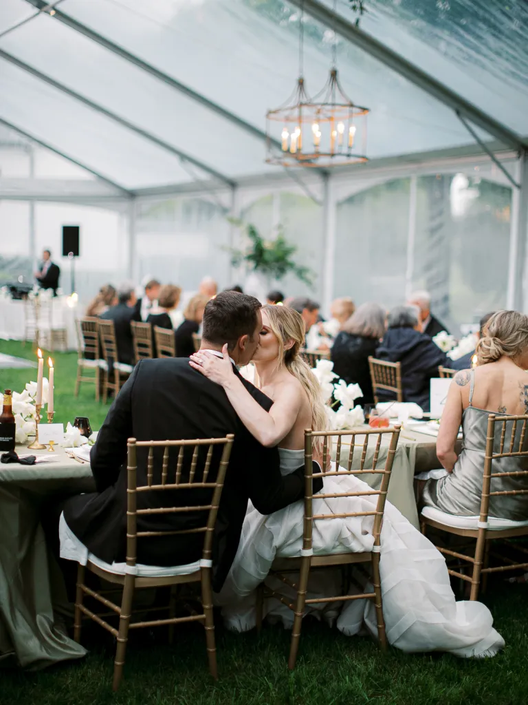Kaitland and Kramer share a romantic kiss at their sweetheart table at Boxwood Estate. Surrounded by elegant floral arrangements and soft candlelight, the couple smiles as they embrace in a moment of joy and love. Kaitland wears a stunning white wedding gown, and Kramer is dressed in a classic suit. The intimate setting, with the estate’s beautiful decor and warm lighting, adds to the magic of their celebration.