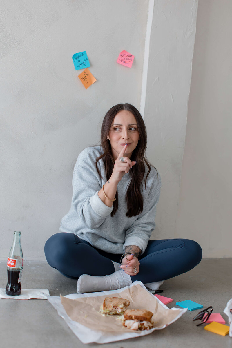 Sharon is sitting on the floor. Looks like lunch time with a sandwich she just took a bite of and her glasses next to her with sticky note pads around.