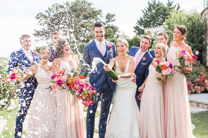 bride pops bottle of champagne with smiling wedding party