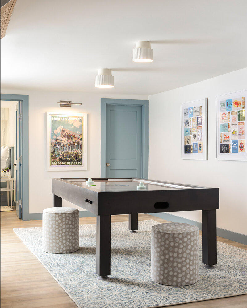 Bright game room featuring a black air hockey table, two patterned round stools, light blue doors, framed art on the walls, and ceiling-mounted lights