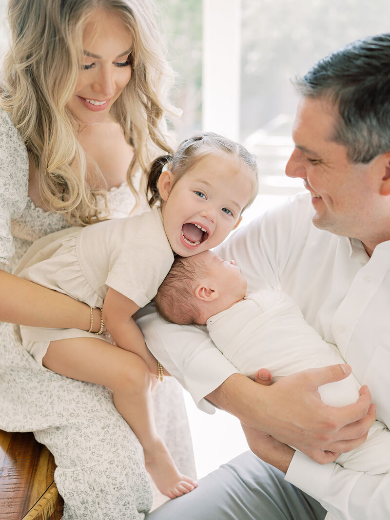 A family of four is gathered together, with a smiling big sister standing beside her parents, who are lovingly holding their swaddled newborn. The family shares a joyful and tender moment, radiating love and excitement for their new addition