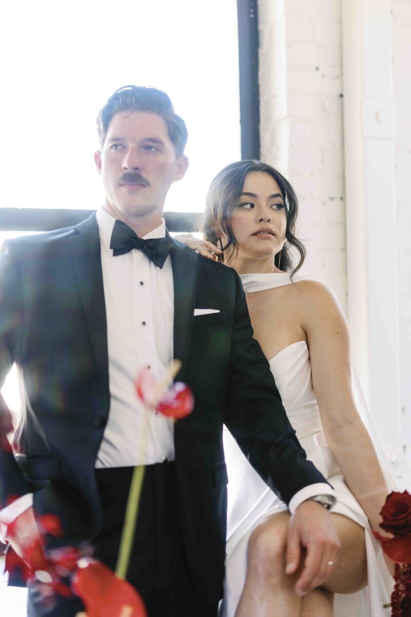 Portrait of bride and groom sitting on window edge with red floral accents.