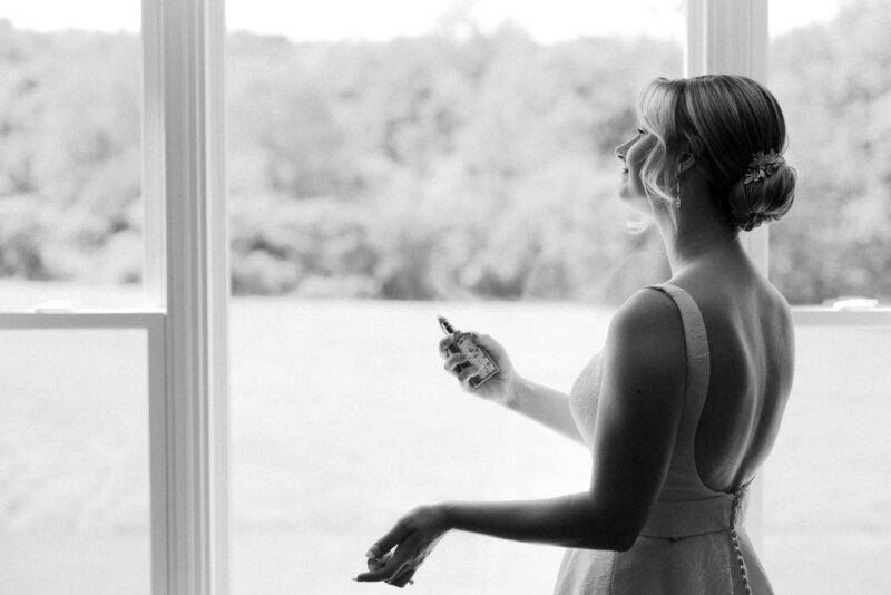 A black and white photo of a bride standing in front of a window spraying her perfume showcasing her bridal hair updo.