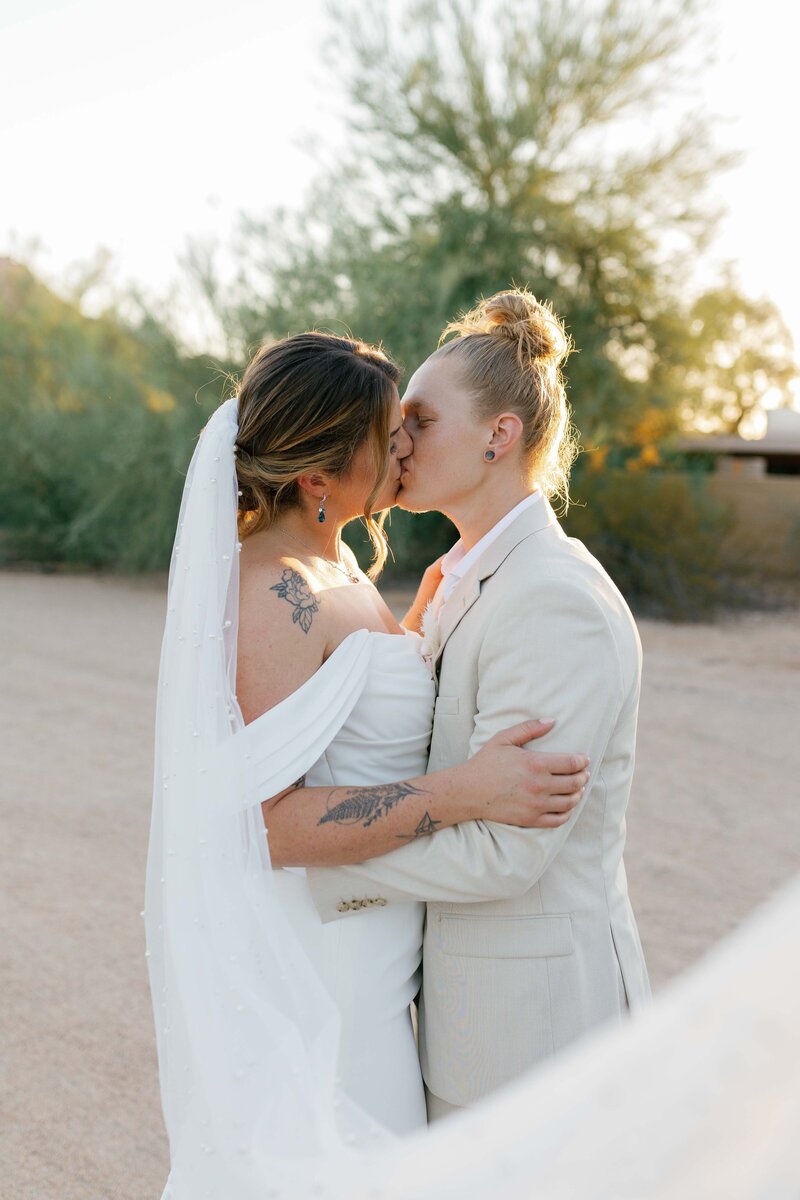 Two brides cuddle and kiss in the desert at sunset.