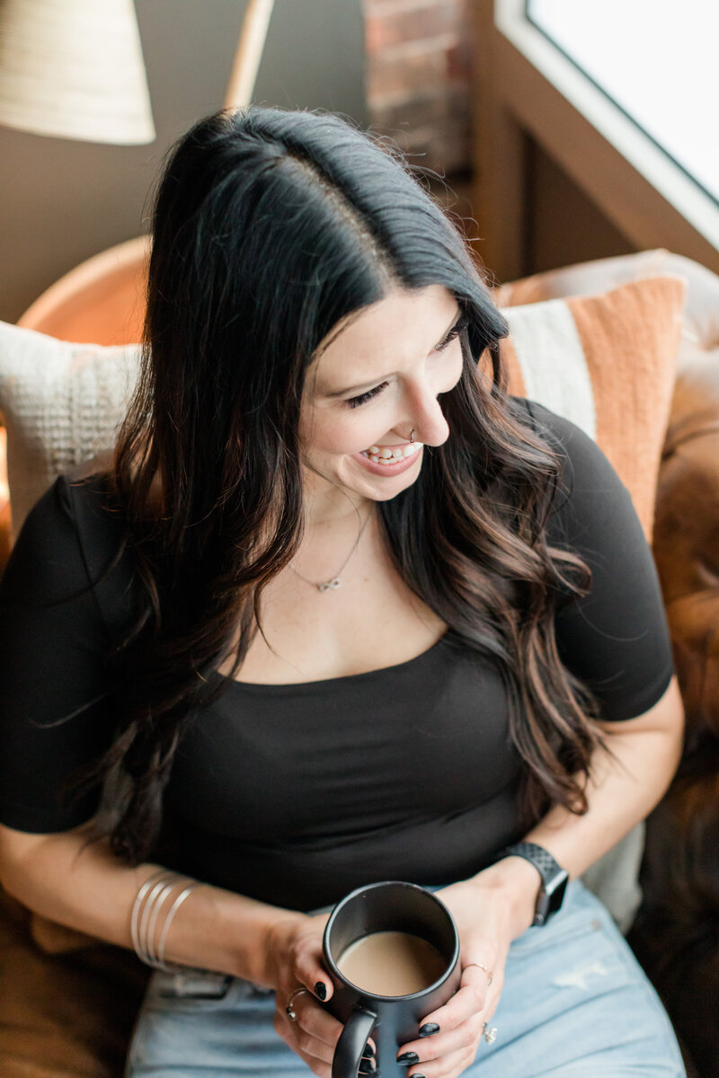 Close-up detail of Allie Clark holding a coffee cup while seated, wearing light ripped jeans and a black top, used to represent the “Design in Progress” phase for Allie Clark Designs branding and website process.