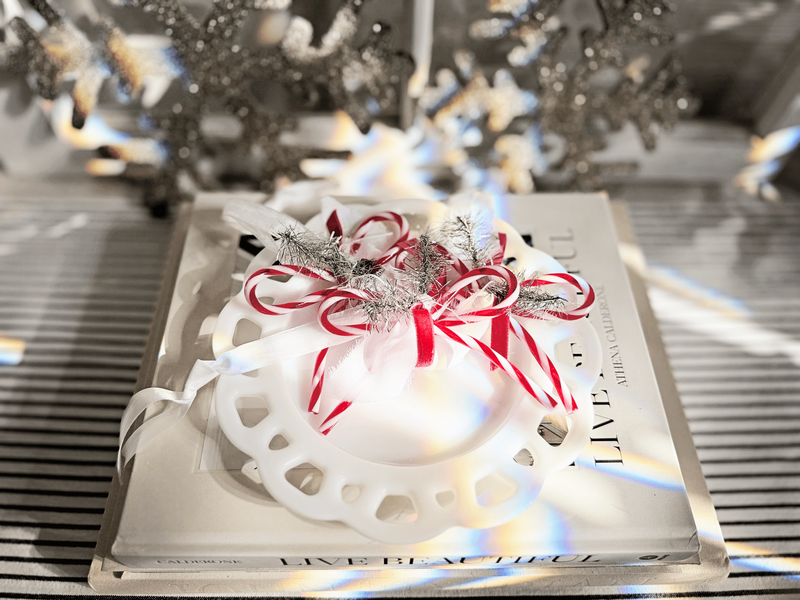 A white decorative plate with red and white ribbon, silver tinsel, and candy cane bow ornaments sits on a book titled "Live Beautiful" by Sweet Pea Ink Creative on a striped surface, with holiday ornaments adding festive cheer in the background.