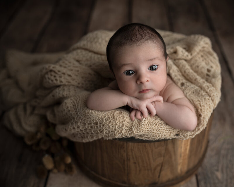 Newborn boy in beige wrap, alert and curious, posed in a rustic bucket for a timeless fine-art portrait.