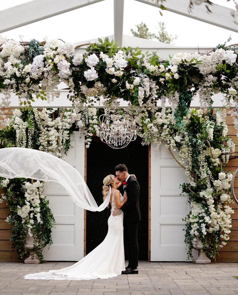 A bride and groom share a kiss under a flower-covered archway at their wedding