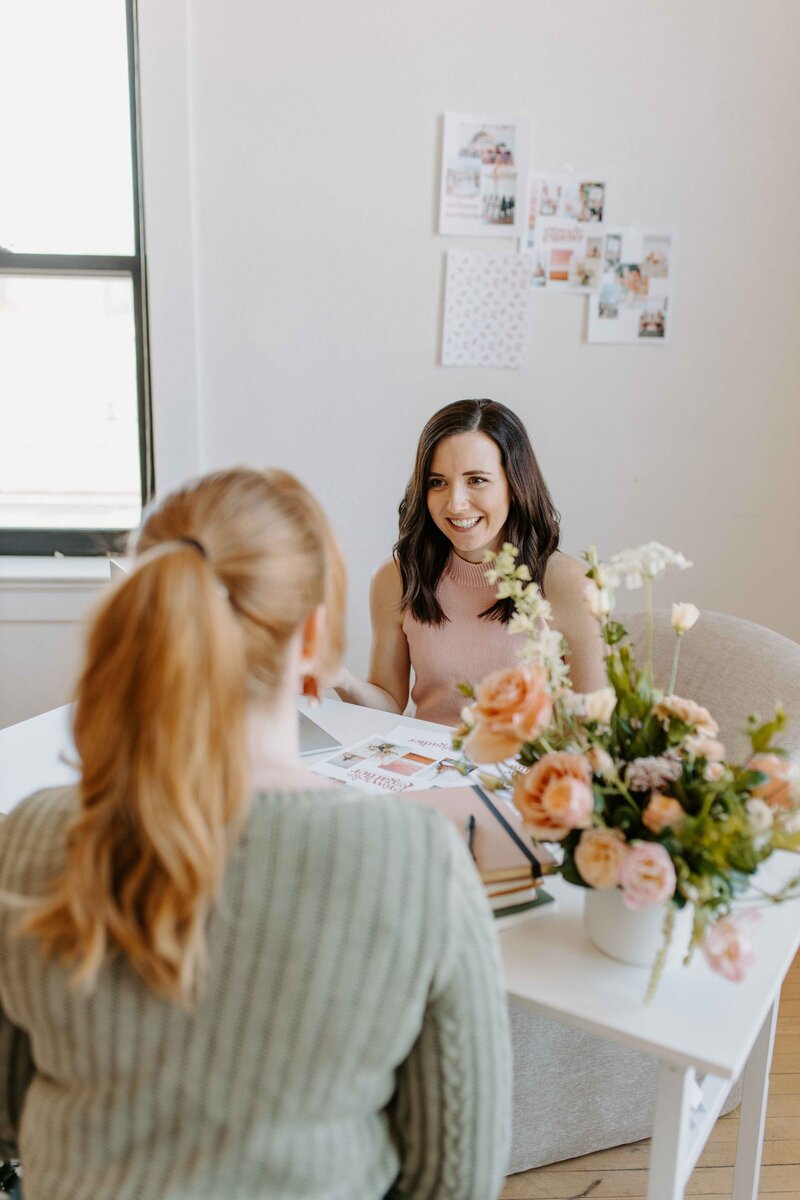 2 women meeting at white desk with flower arangement, one with dark hair and one with strawberry blond hair