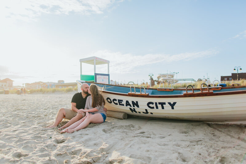 A couple leans against a boat that says Ocean City, NJ, on the beach.
