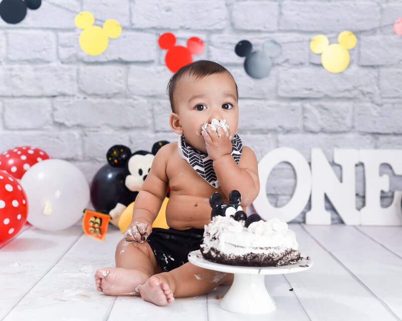 A baby boy enjoying a Mickey Mouse-themed cake smash, with red and white balloons, a "ONE" sign, and a Mickey-inspired backdrop.