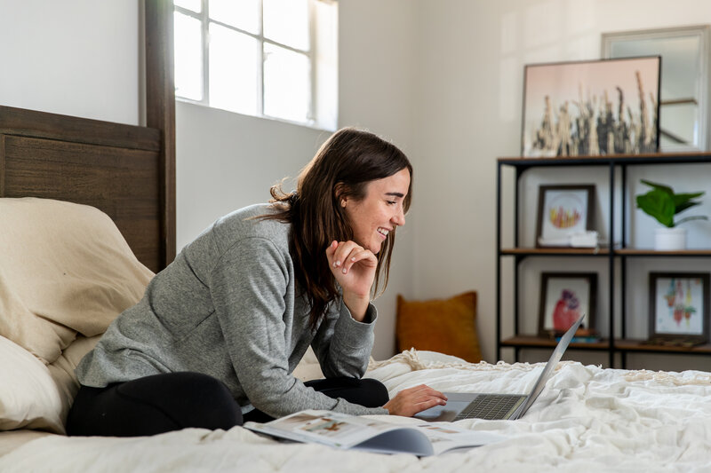 Young woman in a gray sweater sitting cross-legged on a bed, focused on her laptop with natural light coming through the window and artwork displayed on shelves in the background.