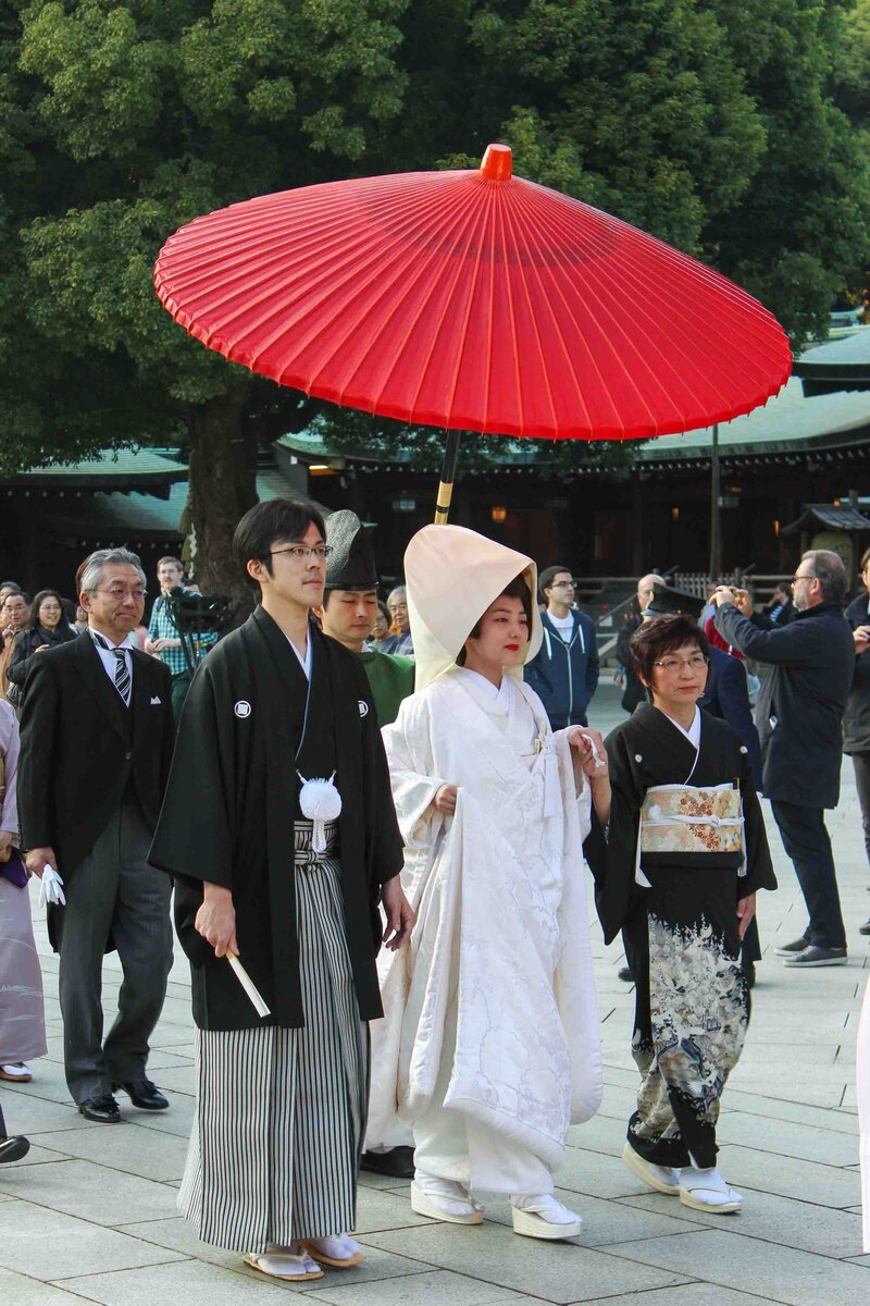 Bride and groom in traditional Shinto wedding attire walking beneath a red paper umbrella at Meiji Shrine in Tokyo ©Stephanie Dosch | theViatrix Japan Private Luxury Travel