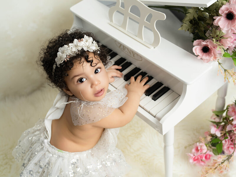 A baby in a mint green lace outfit sits on a small white tufted sofa, with a floral headband and pink flowers nearby, in a soft, serene setting.