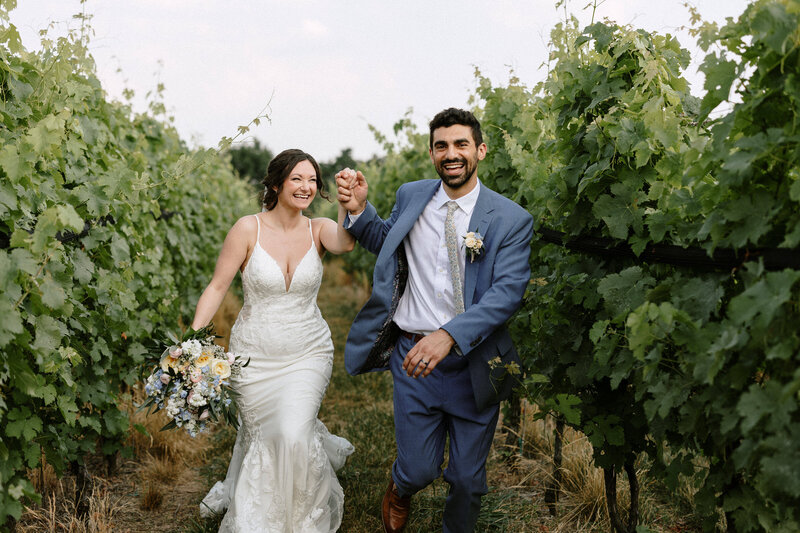 A joyful couple runs hand in hand through a lush vineyard, surrounded by green grapevines, celebrating their wedding day.