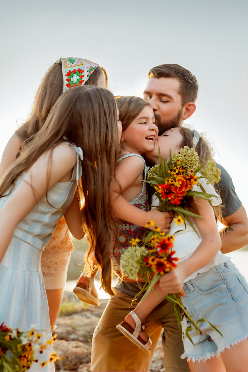 Little girl gets picked up by Dad and gets kisses from mom and two sisters with Kansas City Lifestyle Photographer.