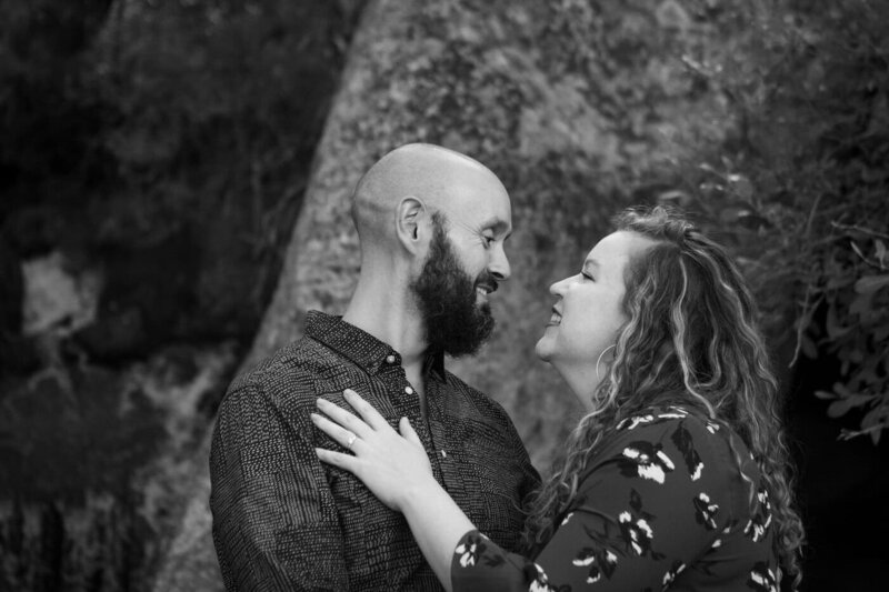 An engaged couple smile at each other  with a simple rock face background.
