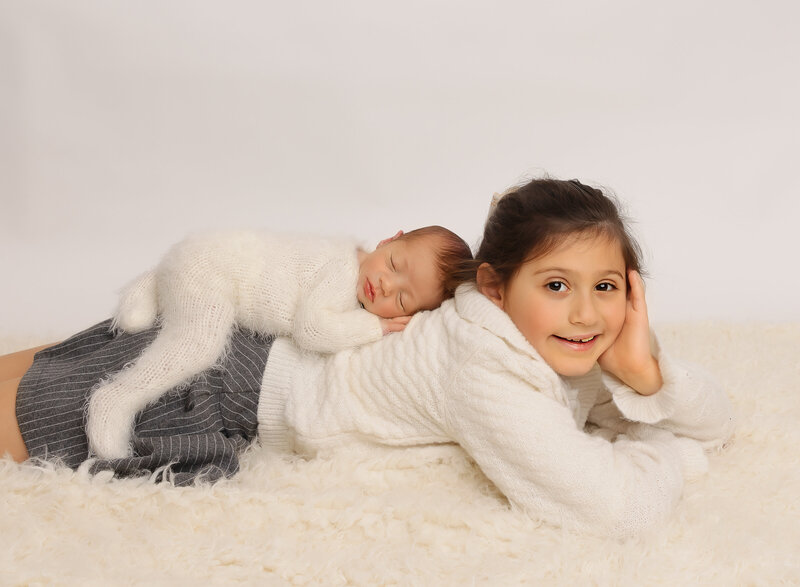 Big sister is lying on her belly side profile to the camera with her head durned toward the camera. She is smiling at the camera. On her back, her new baby brother is laying with his hands underneath his cheek. Captured by premier Brooklyn NY family photographer Chaya Bornstein Photography.
