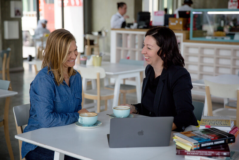 Two women sitting at a table in a cafe, engaged in a conversation about educational leadership. They have coffee cups in front of them and an open laptop on the table, suggesting an academic research session. Books are stacked on a nearby chair, adding to the scholarly atmosphere.
