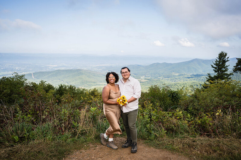 A couple on their elopement day pose together at an iconic viewpoint at Jewel Hollow Overlook in Shenandoah National Park. In the background, there is lots of green foliage as well as the Blue Ridge Mountains.