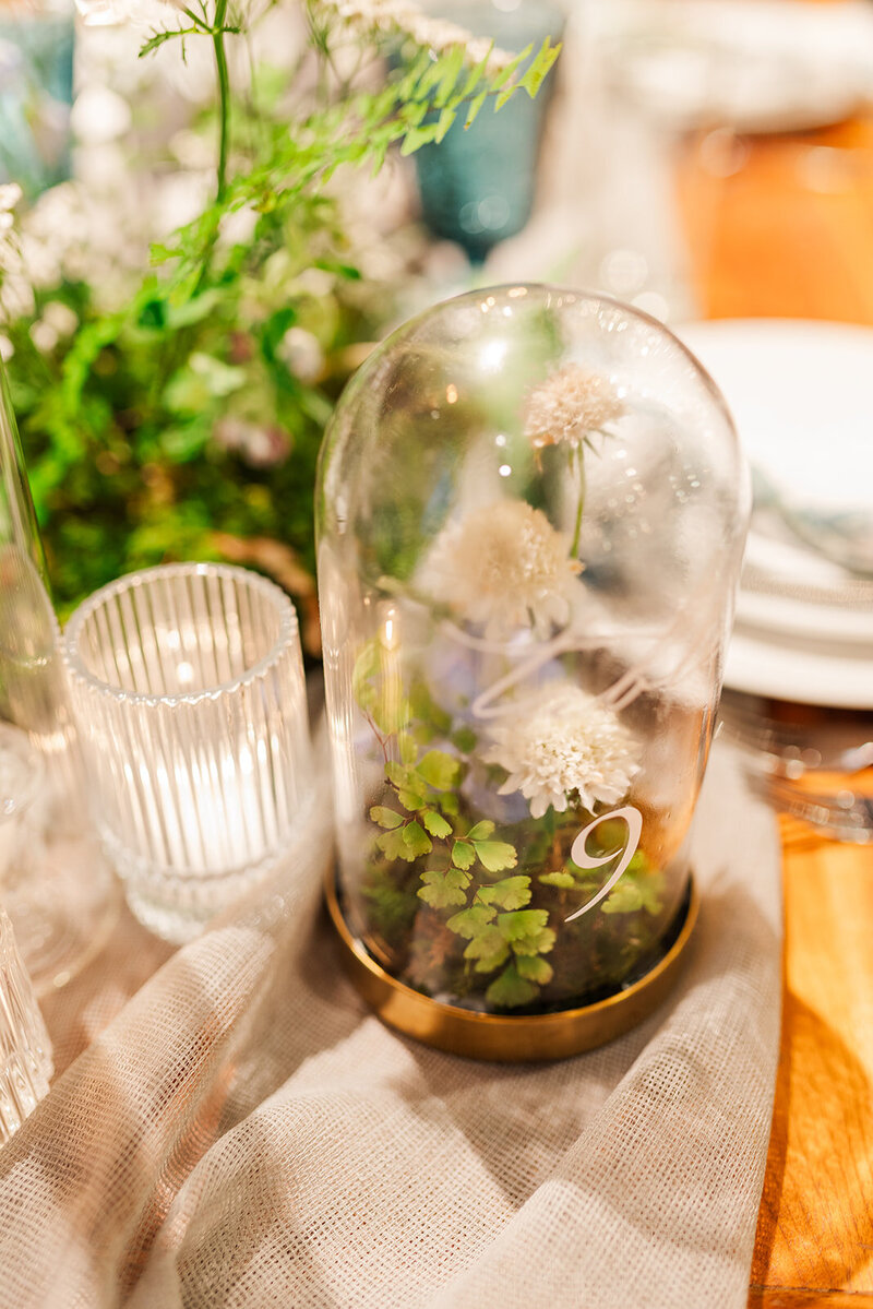 a table number cloche filled with moss and ivory florals  next to a clear ribbed lit votive and moss meadow arrangement on a farm table with a slate colored table runner