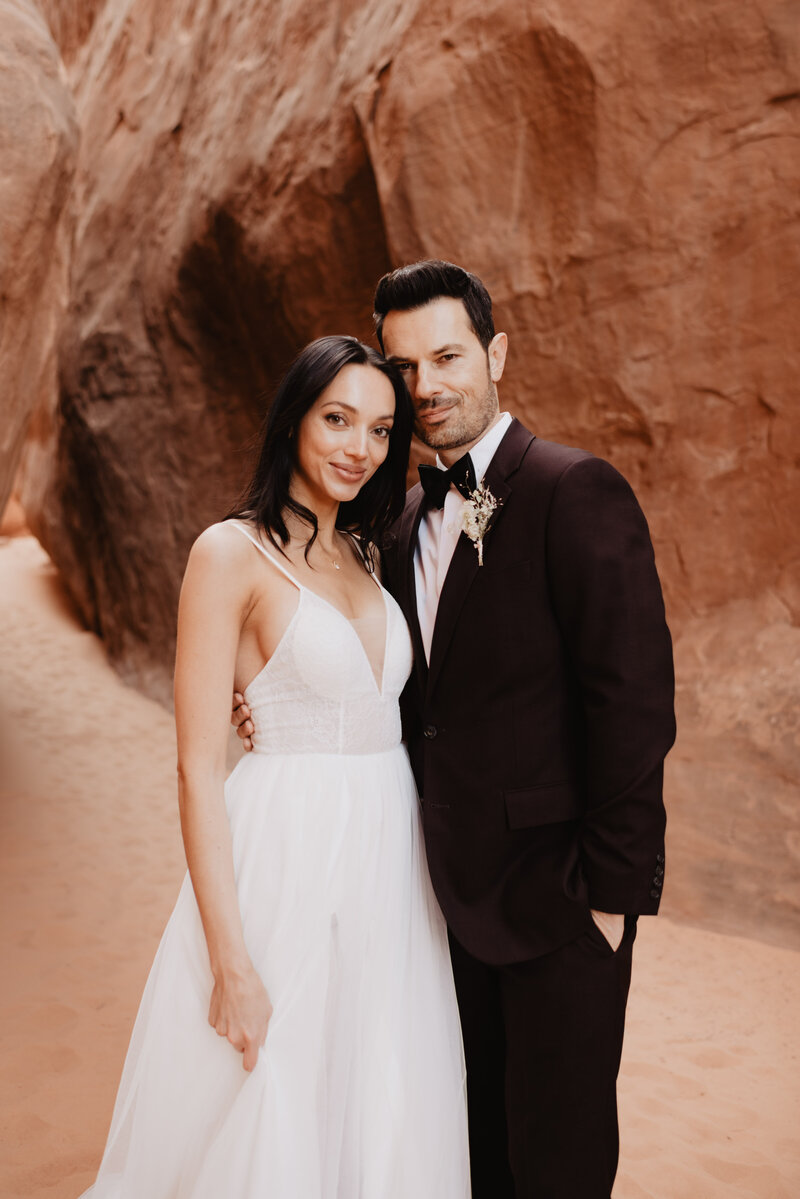 utah elopement photographer captures bridal portrait with groom holding his bride as they both smile in the red rocks of arches national park