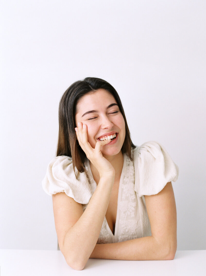 Woman in white blouse smiles while chewing her finger