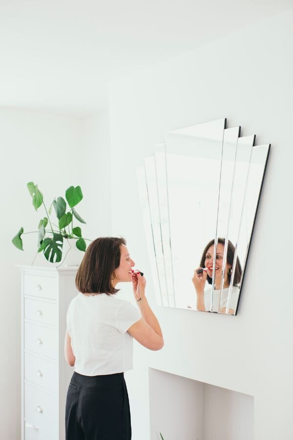 Kaya applying red lipstick while looking into a multi-panel mirror in a bright white room.