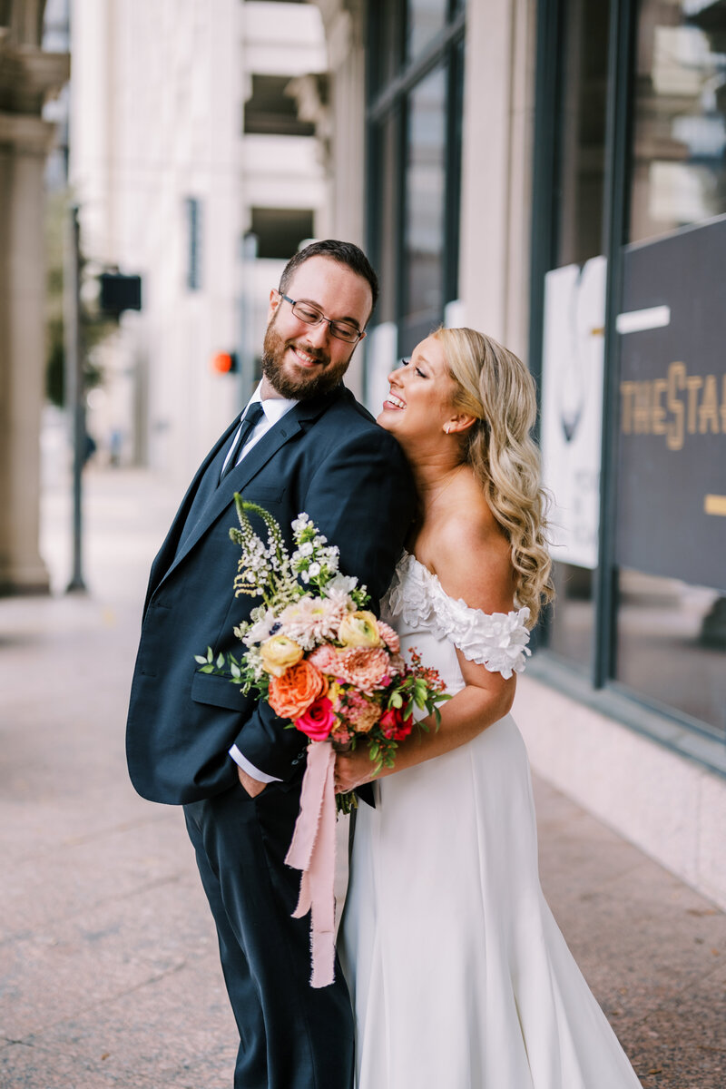 Portrait of bride holding a colorful bouquet and hugging groom from behind while they smile at each other