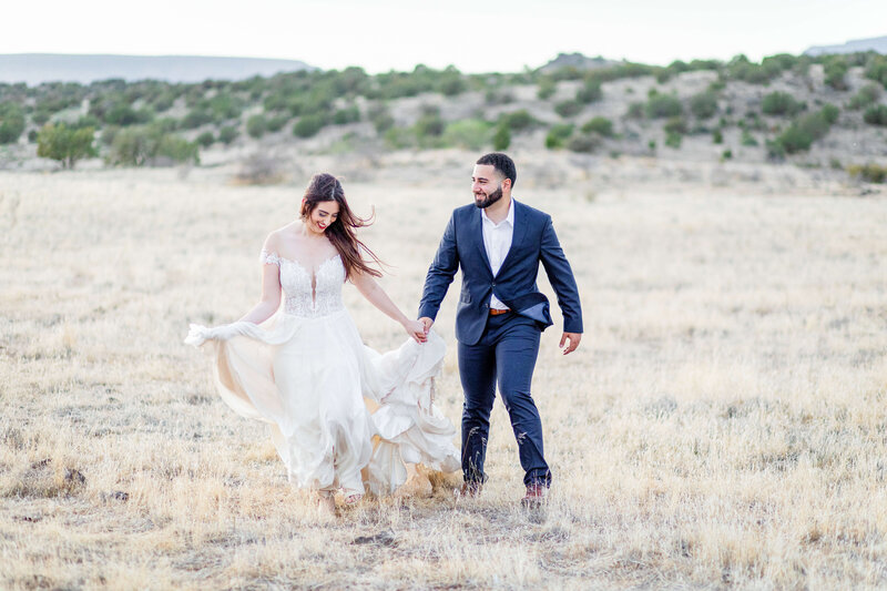 A bride and groom run through an open field with a golden glow.