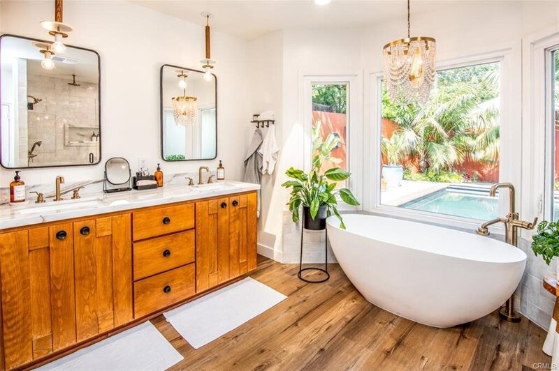 Bright master bathroom with soaking tub, bay window, chandelier, hardwood floors, white walls, and potted ferns for a serene, elevated feel.