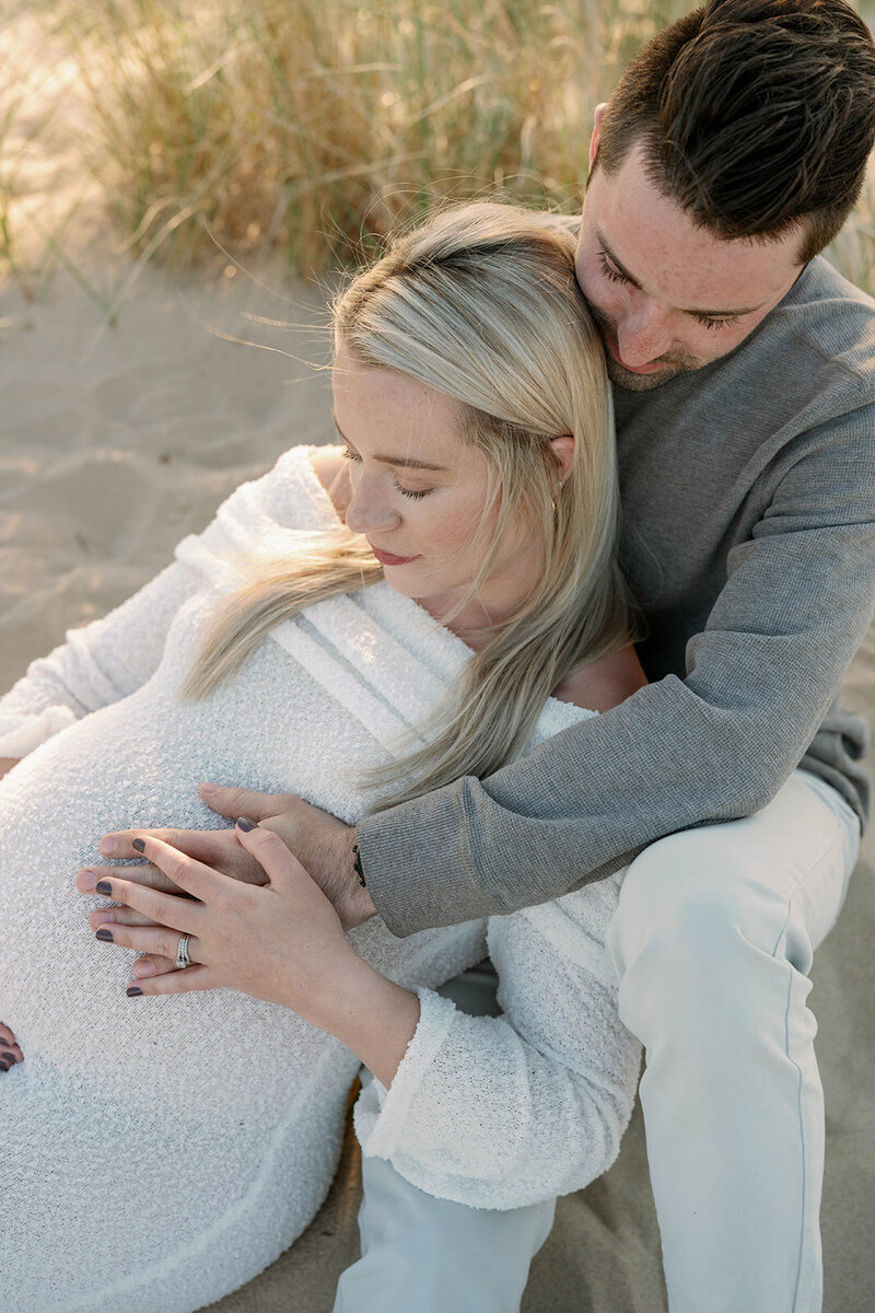 Pregnant woman laying in her husband’s lap while both hold her baby bump during an intimate maternity session.