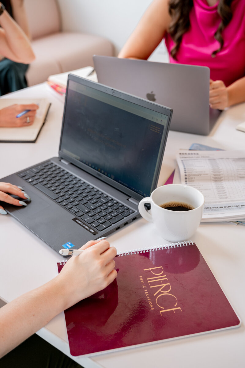 Three Pierce PR team members collaborating around a white table with laptops, notebooks, and coffee mugs, in front of large industrial-style windows.