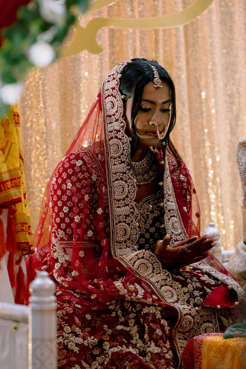 Bride in red and gold lehenga with intricate embroidery, sitting during a traditional Hindu wedding ceremony in New York City, wearing gold jewelry and headpiece