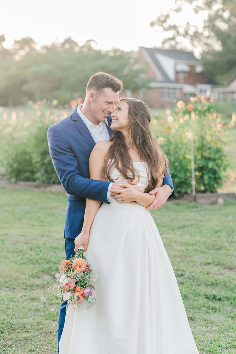 Bride and groom standing in a green field holding each other while bride looks back at husband and holding bouquet