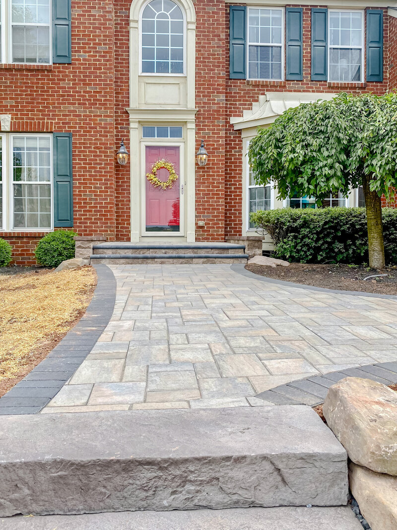 A paver patio with a built in square fire pit. A sitting wall behind the fire pit with green bushes and  a wooden fence behind the sitting wall