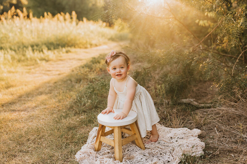 baby girl standing on a blanket and leaning on a little stool in a field at sunset