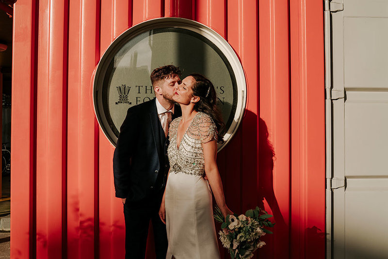 groom kissing bride on a cheek at their trinity buoy wharf wedding with red sea container in the background