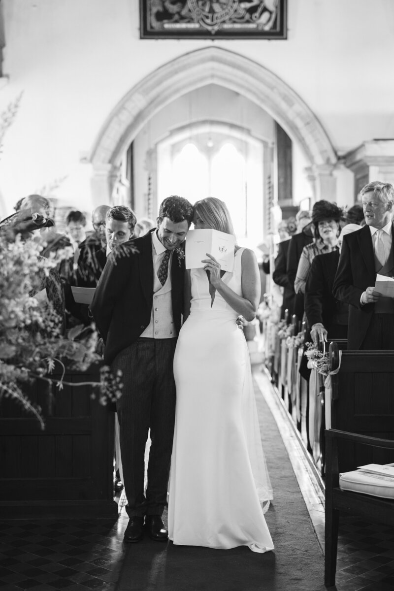 A black and white photo captures a couple at the altar in St Andrew's Church. The Bride, in a long dress, holds a program and leans into the Groom to discretely whisper. Seated guests fill the pews under the grandeur of Winchester's iconic arched window in the background.