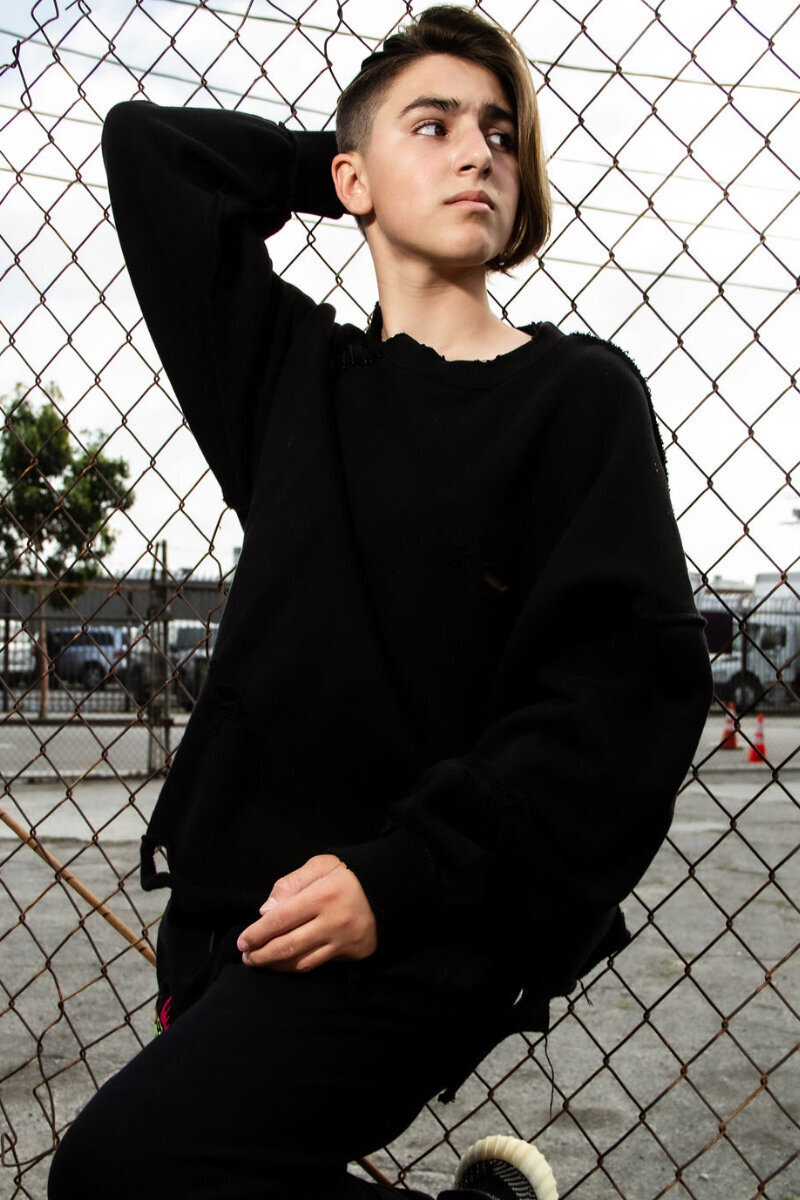 preteen branding portrait young boy standing against chain link fence wearing black shirt and pants one knee bent one arm behind his head